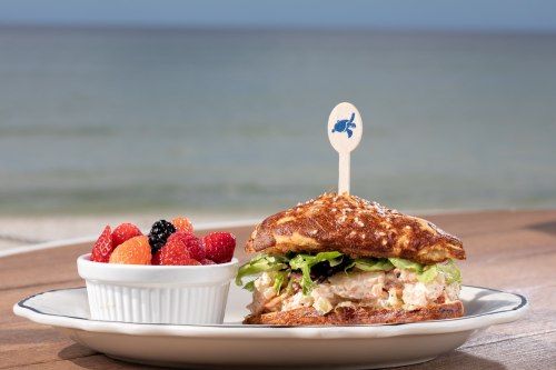 A sandwich with chicken salad and lettuce, served with a side of mixed berries on a wooden table by the beach.
