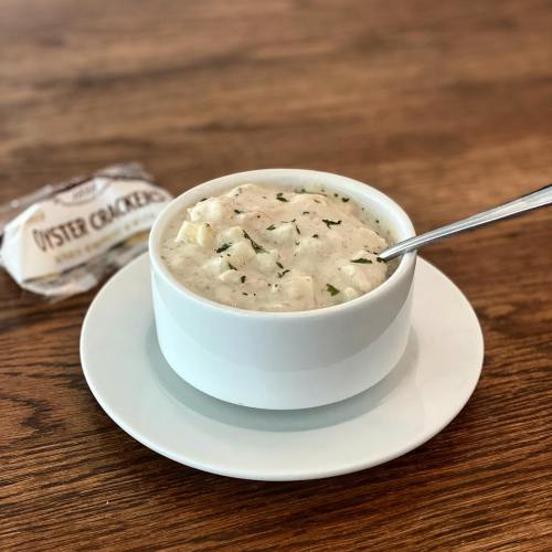 A bowl of creamy soup with herbs, served on a white plate, alongside a pack of oyster crackers on a wooden table.