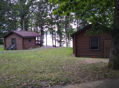 Two rustic cabins surrounded by trees, with a grassy area and bicycles parked nearby, near a lake in the background.