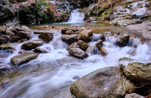 A serene waterfall cascades over rocks into a tranquil stream, surrounded by lush greenery and autumn leaves.