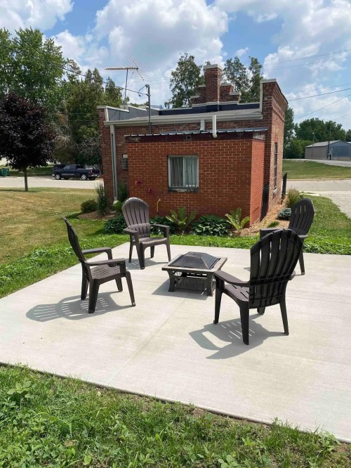 A small brick building with a patio area featuring four black chairs and a table, surrounded by grass and trees.
