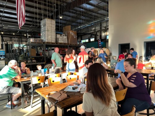A lively gathering in a spacious venue with people socializing at tables and barrels in the background.
