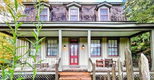 A charming two-story house with a red door, surrounded by greenery and wooden fences, featuring a cozy porch.