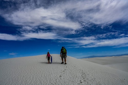 A father and his little girl are hiking in white sand dunes with a bright blue sky overhead.