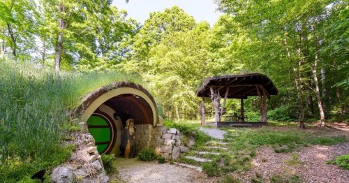 A whimsical hobbit-style home with a grassy roof, surrounded by trees and a nearby rustic gazebo.