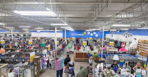 A spacious thrift store filled with clothing racks and shoppers browsing various items. Brightly lit and organized layout.