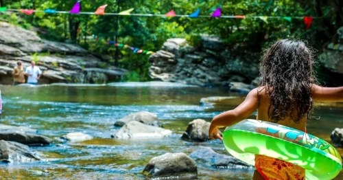 A child with a green inflatable ring walks towards a river, surrounded by rocks and colorful flags in a lush, green setting.
