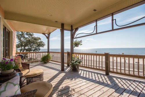 A sunny porch with wicker furniture overlooks a calm beach and water, adorned with potted flowers.
