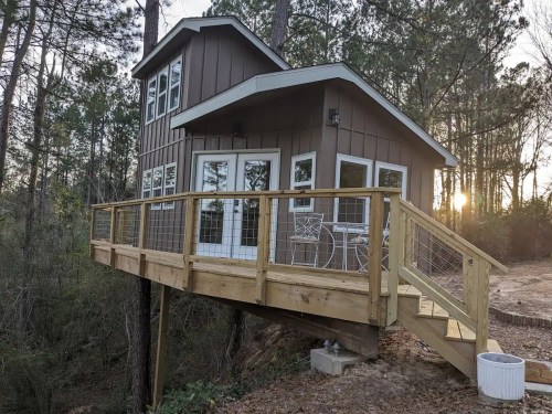 A small, modern cabin on stilts surrounded by trees, featuring a wooden deck and large windows.