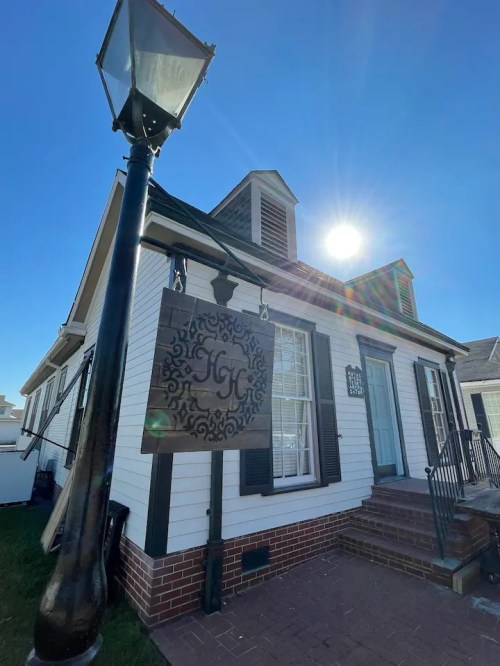 A charming white house with black shutters and a decorative sign, under a bright blue sky and shining sun.