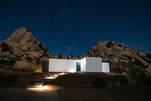 A modern white house illuminated at night, surrounded by rocky terrain and a starry sky.