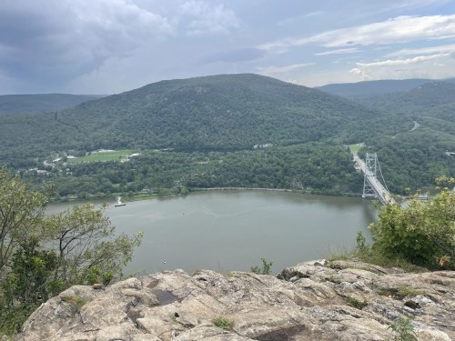 A scenic view of a river winding through green hills, with a bridge crossing over and a cloudy sky above.