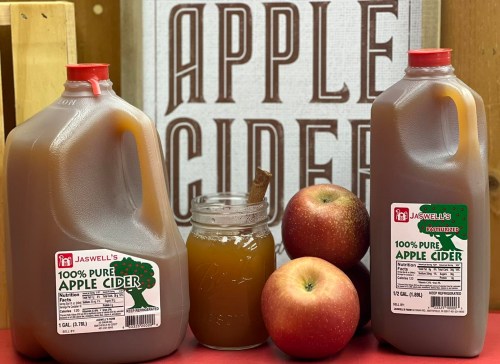 Two jugs of apple cider, a glass of cider, and fresh apples in front of a sign that reads "100% Pure Apple Cider."