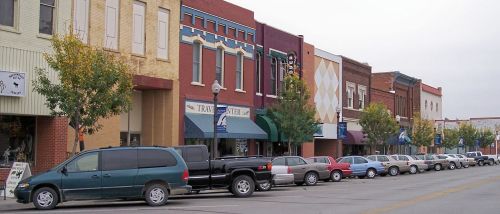 A street view of a small town with parked cars and colorful storefronts along the sidewalk.