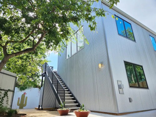 A modern, corrugated metal building with large windows and a staircase, surrounded by greenery and potted plants.