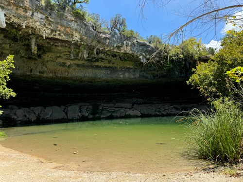A serene natural scene featuring a calm green pool surrounded by rocky cliffs and lush vegetation under a clear blue sky.