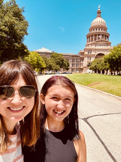 A woman and a girl smile for a selfie in front of a large historic building on a sunny day.