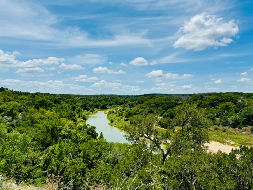 Lush green landscape with a winding river under a bright blue sky dotted with fluffy white clouds.