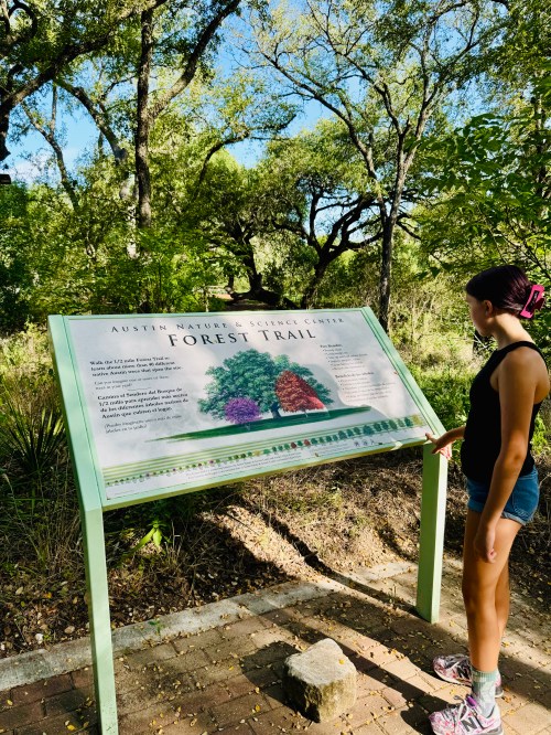 A person stands by a sign for the Forest Trail at the Austin Nature & Science Center, surrounded by greenery.