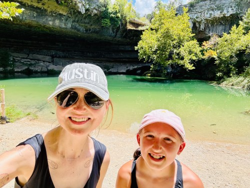 A woman and a girl smile by a green lake surrounded by trees and rocky cliffs on a sunny day.