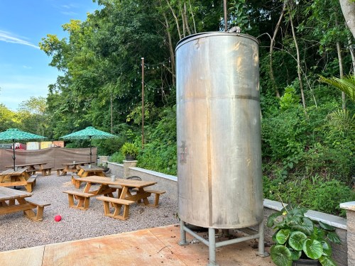 A large silver tank stands beside picnic tables in a gravel area, surrounded by greenery and shaded by umbrellas.