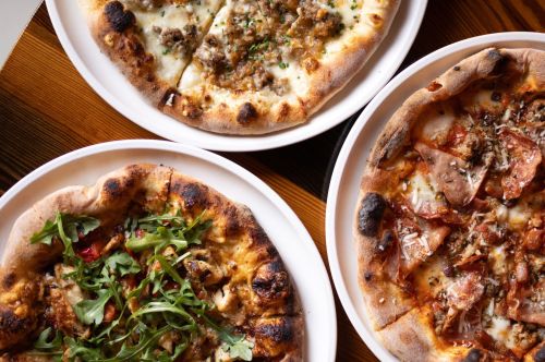 Three pizzas on white plates: one with greens, one with meat, and one with cheese and toppings, on a wooden table.