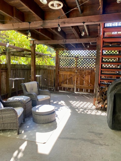 Cozy outdoor patio with wicker chairs, a round table, and wooden beams, illuminated by string lights and surrounded by greenery.