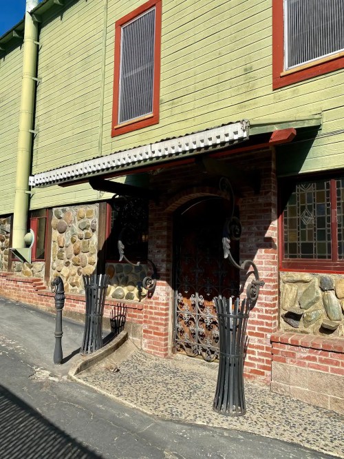 A green building with a stone facade, featuring an ornate iron gate and decorative fencing along a cobblestone path.