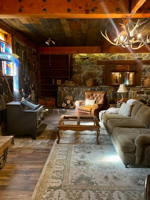Cozy rustic living room with stone walls, a leather armchair, a sofa, and a wood stove, illuminated by natural light.