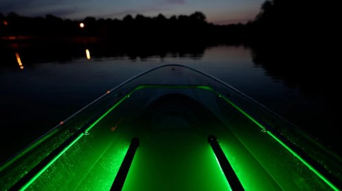 A glowing green-lit boat on calm water at dusk, with trees and lights reflected in the background.