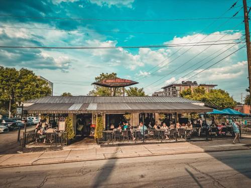 A lively restaurant with outdoor seating, filled with diners, under a bright blue sky and scattered clouds.