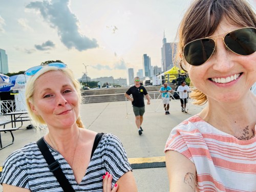 Two women smile for a selfie outdoors, with a city skyline in the background and a man walking behind them.