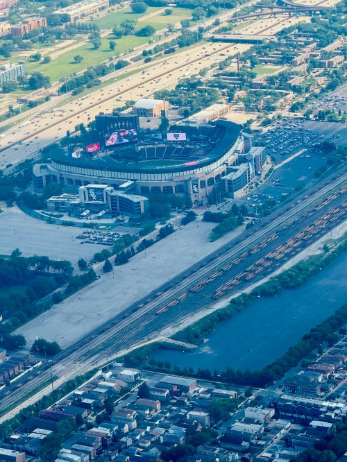 Aerial view of a stadium surrounded by parking lots, train tracks, and urban landscape. Green fields in the background.