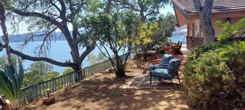 A serene lakeside view with chairs under trees, surrounded by greenery and a clear blue sky.