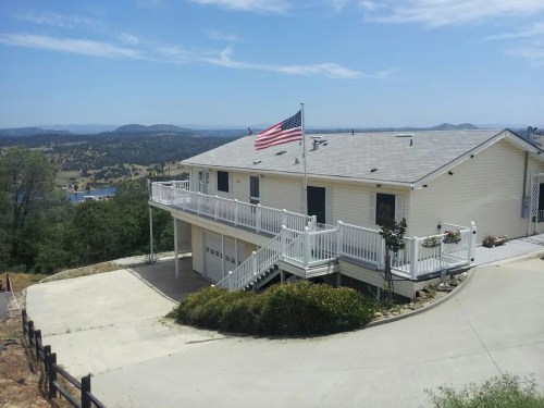 A two-story house on a hillside with a U.S. flag, surrounded by greenery and mountains in the background.