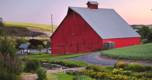 A vibrant red barn stands beside a lush garden, with rolling hills in the background under a pastel sky.