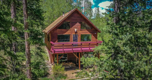 A wooden cabin surrounded by tall trees, featuring a balcony and large windows, set in a lush green landscape.