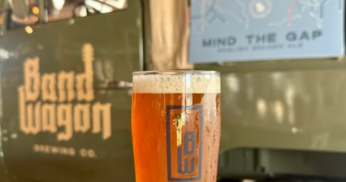 A glass of amber beer in front of a vintage brewing truck with "Band Wagon Brewing Co." logo.
