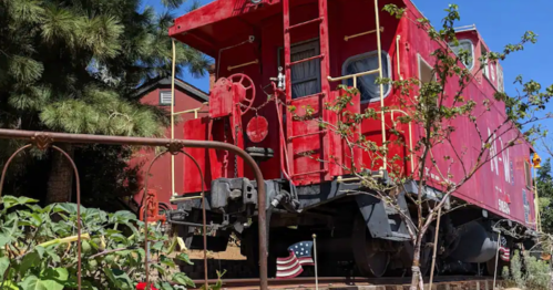 A red caboose on display, surrounded by greenery and a small American flag in the background.