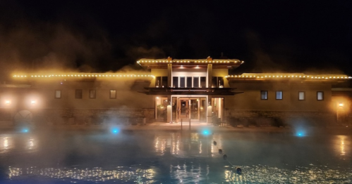 A serene spa building at night, illuminated with warm lights, surrounded by mist rising from a tranquil pool.