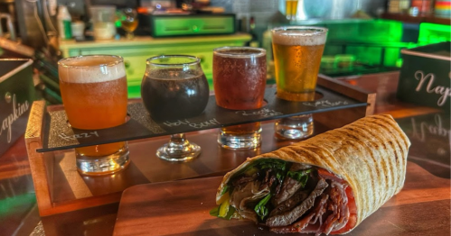 A wooden platter with four beer samples and a wrap filled with greens and meat in the foreground.