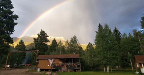 A rainbow arcs over a rustic cabin surrounded by trees and mountains under a cloudy sky.
