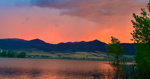A serene lake at sunset, with vibrant orange and pink skies reflecting on the water and mountains in the background.