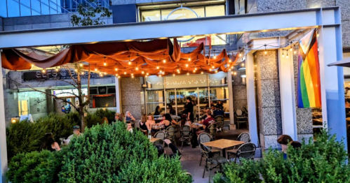 Outdoor dining area with string lights, patrons at tables, and a rainbow flag hanging nearby.