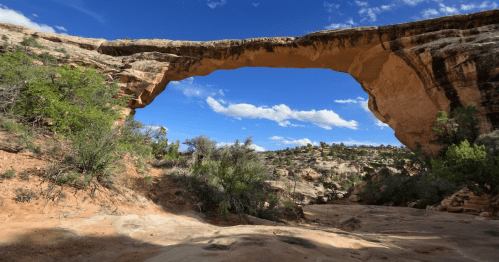Natural stone arch against a blue sky with clouds, surrounded by desert vegetation and rocky terrain.