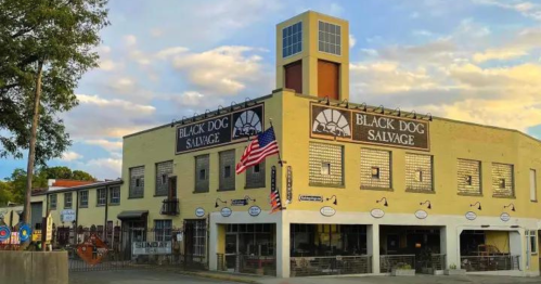 A yellow building with "Black Dog Salvage" signage, featuring large windows and an American flag in front.