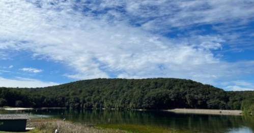 A serene landscape featuring a calm lake surrounded by lush green hills and a blue sky with scattered clouds.