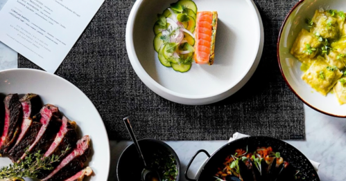 A table spread featuring sliced steak, sushi, cucumber salad, and dumplings on a textured placemat.