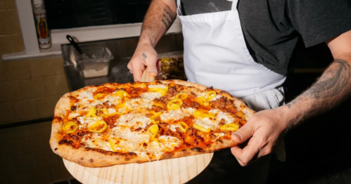 A person in an apron holds a large pizza topped with cheese and yellow peppers, ready to serve.