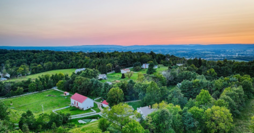 Aerial view of a lush green landscape with houses and a red-roofed barn at sunset, surrounded by rolling hills.